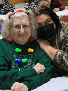 Two women in holiday attire smiling at camera. Elderly woman has antlers hat on and Christmas lights around her neck.